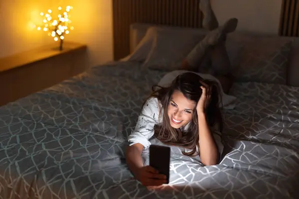 Neşeli Genç Bir Kadın Geceleri Yatakta Uzanıyor Akıllı Telefon Kullanarak — Stok fotoğraf
