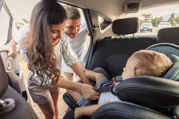 Parents fastening cute little toddler son into a baby car seat, getting ready for safe drive with their child