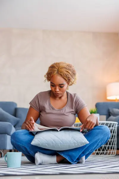 リビングフロアに座り 雑誌を読み 自宅でリラックスしながらコーヒーを飲んでいる美しいアフリカの女性 — ストック写真
