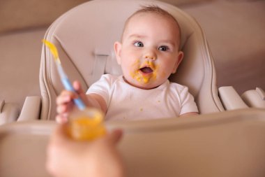Neşeli erkek bebek yüksek sandalyede tek başına oturmuş yulaf lapası yerken gülüyor.