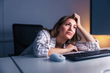 Ofiste geç saatlere kadar çalışan yorgun ve stresli bir kadın masasına yaslanmış ve masaüstü bilgisayar ekranına bakarken elini tutuyor.