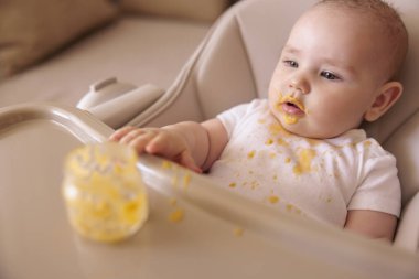 Küçük sevimli erkek bebek, lapa yedikten sonra dağınık ve lekeli bir şekilde yüksek sandalyede oturuyor.