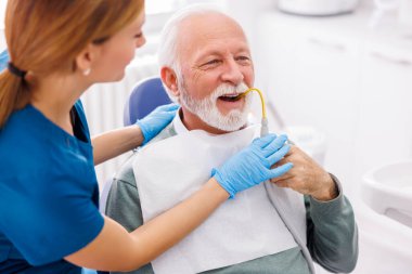 Dişçi koltuğunda oturan kıdemli adam elinde tükürük emme tüpü tutarken doktor dişini düzeltiyor.
