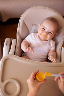 Annesinin neşeli erkek çocuğunu lapayla beslediği yüksek açılı manzara, bebek sandalyede oturuyor ve gülümsüyor ve yemek yiyor.