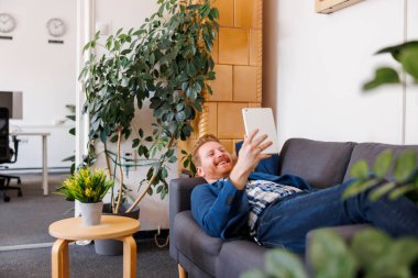 Ofisteki kanepede uzanmış, tablet bilgisayar kullanarak video görüşmesi yapan genç bir iş adamı.