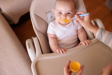 Annesinin neşeli erkek çocuğunu lapayla beslediği yüksek açılı manzara, bebek sandalyede oturuyor ve gülümsüyor ve yemek yiyor.