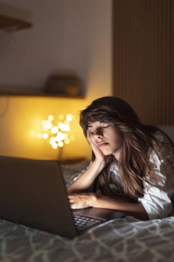 Güzel bir kadın yatakta yatıp fazla mesai yapıyor. Dizüstü bilgisayar kullanıyor, yorgun ve uykulu.