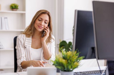 Sıradan bir iş kadını telefon görüşmesi yaparken akıllı telefon kullanır ve ofiste çalışırken planlamacıdan not alır.