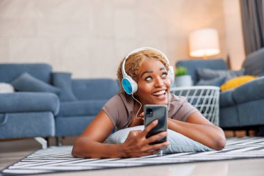 Kulaklık takmış bir kadın evde dinlenirken akıllı telefon kullanarak internette sörf yapıyor ve boş zamanların tadını çıkarıyor.