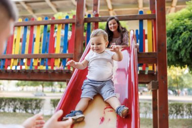 Anne ve baba parkta kaydırağın üzerinde oynaşıp eğleniyorlar.