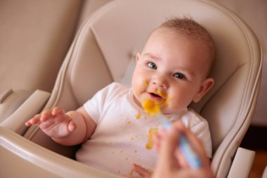 Anne, neşeli erkek çocuğu lapa ile besliyor, bebek, yüksek sandalyede oturuyor, dağınık oturuyor, gülümsüyor ve yemek yiyor.