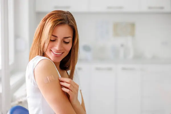 Portrait of young woman sitting in healthcare facility with medical patch on her arm after vaccination - vaccination, immunization and disease prevention concept
