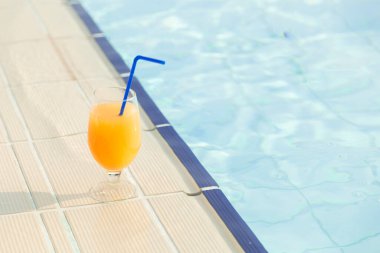 Güneşli bir yaz gününde yüzme havuzunun yanına konmuş bir bardak portakal suyu ve pipet.