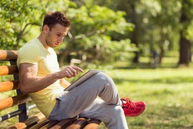 Sıkı bir antrenmandan sonra genç sporcu, parktaki ahşap bir bankta oturuyor, tablet bilgisayarda internette sörf yapıyor.