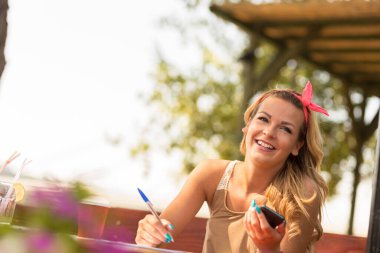 Güzel, sarışın bir kız açık bir kafede oturuyor, telefonla konuşuyor ve notlar yazıyor.