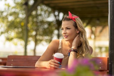 Güzel sarışın bir kız açık hava kafede oturuyor, elinde bir fincan kahve tutuyor ve derin derin uzaklığa bakıyor.