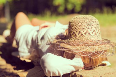 Güneşli bir yaz gününde, bir parkta tuğla duvarda uzanmış, şapka takmış genç bir kız.