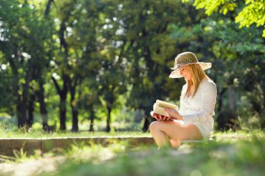 Parkta güneşli bir yaz gününde kitap okuyan genç bir kadın.