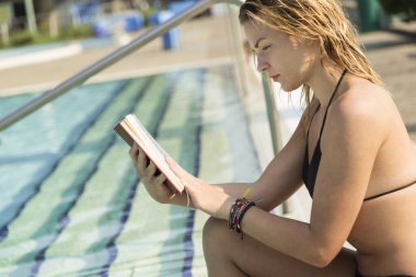 Genç, güzel sarışın kız yüzme havuzunun yanında oturuyor ve kitap okuyor.