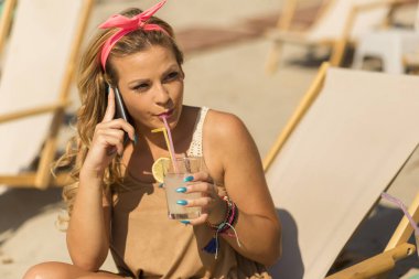 Kumsalda güneşli bir yatakta oturup limonata içen ve telefonda konuşan güzel sarışın kız.