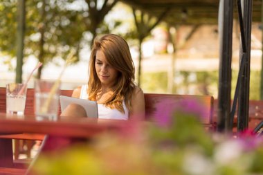 Güzel bir esmer açık hava kafede oturuyor, tablet bilgisayarında e-kitap okuyor ve limonata içiyor.