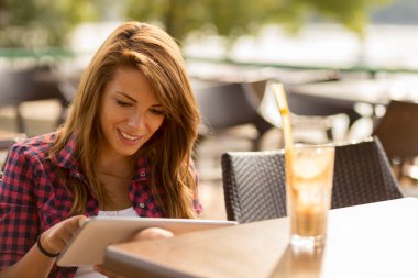 Güzel bir esmer açık hava kafede oturuyor, tablet bilgisayarında e-kitap okuyor ve limonata içiyor.