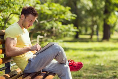 Sıkı bir antrenmandan sonra genç sporcu, parktaki ahşap bir bankta oturuyor, tablet bilgisayarda internette sörf yapıyor.