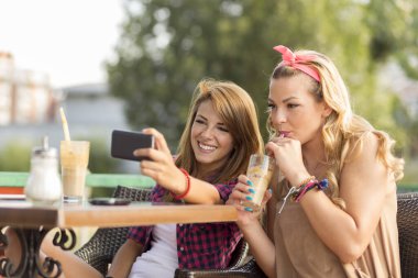 Güzel genç kızlar açık bir kafede oturuyor, kahve içiyor ve birbirlerinin fotoğraflarını çekerken eğleniyorlar.