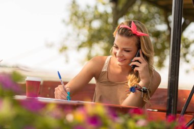 Güzel, çekici, sarışın bir kız bir kafede oturuyor, elinde cep telefonu, ve notlar yazıyor.