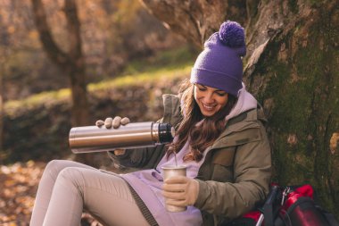 Aktif genç bir kadın ormanda oturuyor, ağaca yaslanıyor, yürüyüş molası veriyor güneşli bir sonbahar gününde dışarıda dinleniyor ve vakumlu bir şişeden bir fincan sıcak çay dolduruyor.