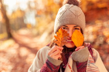 Neşeli genç bir kadın güneşli bir sonbahar gününü doğada geçirip renkli yapraklarla arkalarına bakarak eğleniyor.