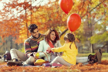 Genç bir aile piknik battaniyesinde oturuyor, sandviç yiyor, eğleniyor ve doğada güzel bir sonbahar gününün tadını çıkarıyor.