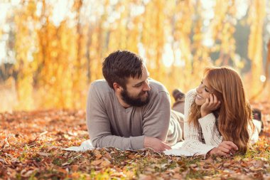 Birbirine aşık genç bir çift, düşen bir sonbahar yaprakının üzerinde parktaki bir ağacın altında yatıyor ve harika bir sonbahar gününün tadını çıkarıyor.