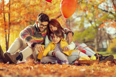 Güzel, genç bir aile piknik battaniyesinde oturuyor, eğleniyor ve parkta sonbahar gününün tadını çıkarıyor.