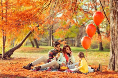 Genç bir aile piknik battaniyesinde oturuyor, sandviç yiyor, eğleniyor ve doğada güzel bir sonbahar gününün tadını çıkarıyor.