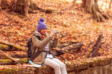 Aktif bir kadın ormanda oturmuş yürüyüş yapıyor, doğanın fotoğraflarını çekiyor ve sonbahar macerasının tadını çıkarıyor.