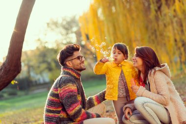 Aile doğada bir sonbahar gününün tadını çıkarıyor, ebeveynler çocuklarıyla oynuyor, küçük kız sabun köpüğü yapıyor.