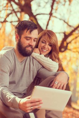 Birbirine aşık bir çift, sonbahar yaprakları üzerinde parkta oturup tablet bilgisayarında internette sörf yapıyor.