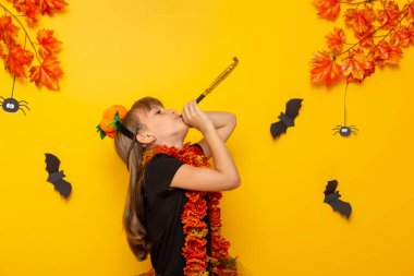 Cadılar Bayramı için parti düdüğü çalan cadı kostümlü çocuk, sarı renkli arka planda izole edilmiş.