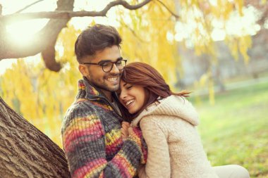 Genç aşık çift ağaca yaslanıyor, sarılıyor ve parkta güneşli bir sonbahar gününün tadını çıkarıyor. Kız arkadaşına odaklan.