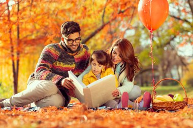 Anne, baba ve kız parkta sonbahar pikniğinde eğleniyorlar, hikaye okuyorlar.