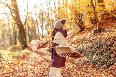 Güzel genç bir kadın, yapraklarla kaplı renkli sonbahar ormanı yolunda yürürken daireler çizerek eğleniyor.