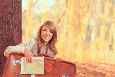 Güzel genç bir kadın, elinde bavul ve haritayla parktaki düşen sonbahar yapraklarında oturuyor ve tatile hazırlanıyor.