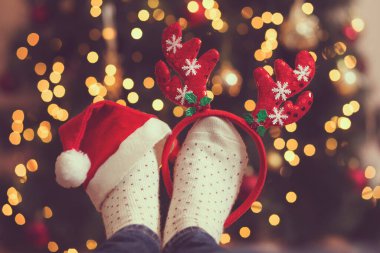 Sıcak kış çorapları, boynuzlar ve Noel Baba şapkası giyen bir kadın. Masaya yerleştirilmiş. Arka planda Noel ağacı ve Noel ışıkları var. Boynuzlara odaklan.