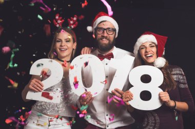 Üç genç arkadaş yeni yıl partisinde eğleniyor, ellerinde 2018 numaralı karton kutular var.