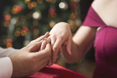 Young attractive couple in love sitting at a dinner table next to a nicely decorated Christmas tree while man surprises his girlfriend with an engagement ring clipart