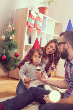 Genç bir aile Noel ağacının yanında oturuyor, parti şapkaları takıyor ve kızlarının doğum günü pastasına mum üflüyorlar. Kızına odaklan.