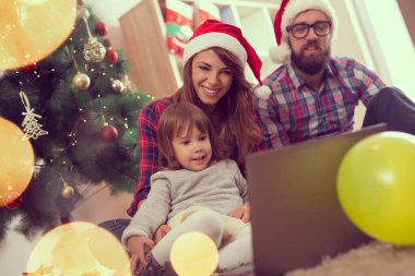 Genç ve güzel bir aile oturma odasının zemininde oturmuş güzel dekore edilmiş bir Noel ağacının yanında dizüstü bilgisayarda çizgi film izliyorlar.