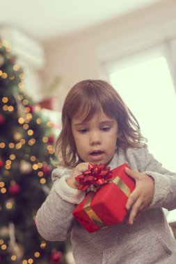 Güzel dekore edilmiş bir Noel ağacının yanında duran küçük kız hediyesini açıyor.