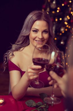 Birbirine aşık bir çift yemek masasında oturmuş, arka planda güzel dekore edilmiş Noel ağacı ve bir kadeh şarapla kadeh kaldırıyor.